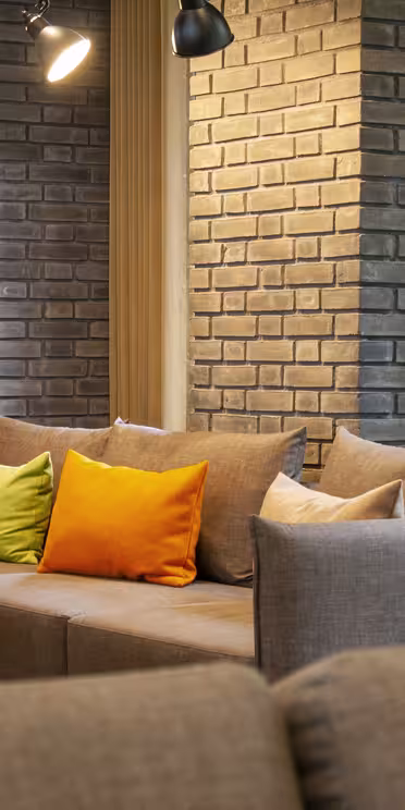 Ett mysigt vardagsrum med en grå sektionssoffa prydd med färgglada orange, gröna och beige kuddar. Rummet har tegelväggar, vertikala träribbor, varm belysning och ett öppet fönster med utsikt över grönskan utanför.