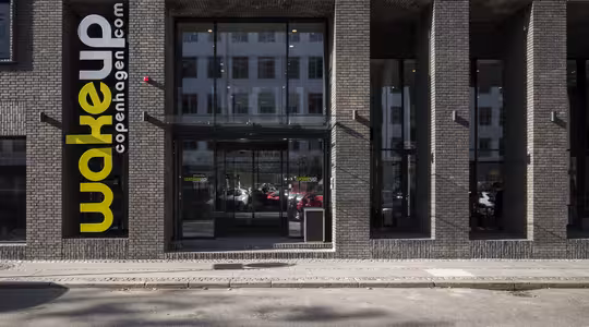 Fasad av en modern hotellbyggnad med mörkt tegel. Stora entrédörrar i glas är centrerade och hotellets namn i gult och vitt på en vertikal svart skylt lyder "wakeup copenhagen.com." Reflexioner av närliggande byggnader syns i glaset.