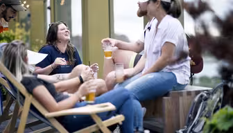 En grupp människor som sitter och står på ett tak och pratar och skrattar. De håller drinkar medan de njuter av en avslappnad atmosfär, med avslappnad klädsel och en urban bakgrund med grönska och dekorativa ljus.
