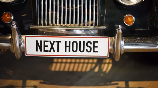 Närbild av en veteranbils front, med en grill, runda strålkastare och en vit registreringsskylt med texten "NEXT HOUSE". Bilen är blank och klassisk i stilen.