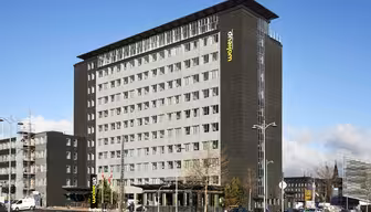 Ett modernt hotell i flera våningar med mörkgrå exteriör och stora fönster, döpt till "Wakeup" med en gul logotyp. Byggnaden är omgiven av en parkeringsplats, gatlyktor och några parkerade bilar. Himlen är klar med några moln.