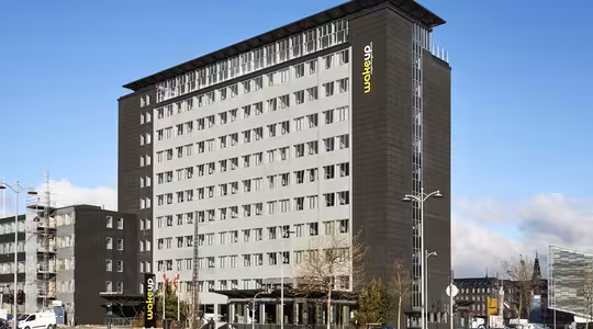 Ett modernt hotell i flera våningar med mörkgrå exteriör och stora fönster, döpt till "Wakeup" med en gul logotyp. Byggnaden är omgiven av en parkeringsplats, gatlyktor och några parkerade bilar. Himlen är klar med några moln.