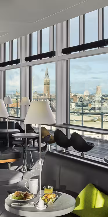 En modern lounge med stora fönster med utsikt över staden. Utrymmet har bord med gröna stolar, svarta soffor och vita lampor. Krukväxter ger grönska, och en frukostmiljö med juice och ett bakverk står på ett bord.