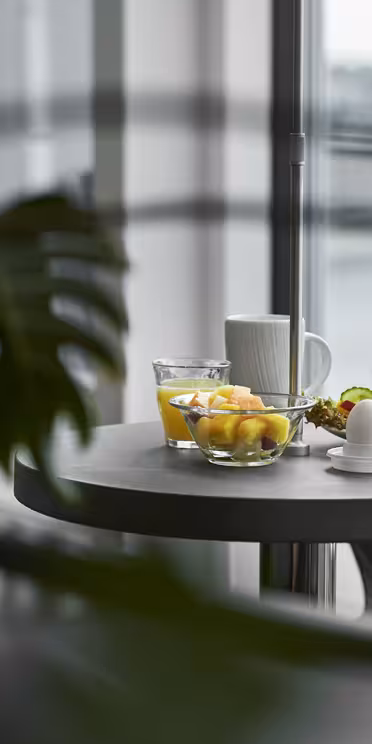 Ett modernt matbord med en skål med frukt, en kopp och en tallrik med ett bakverk och ett litet fat, delvis skymd av gröna växtblad. Bakgrunden har stora fönster med suddig utsikt utanför.