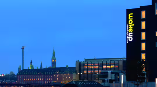 Stadsbild i skymningen med utsikt över stadsbyggnader och en upplyst skylt med texten "vakna upp" på en mörk byggnad. Skyline har ett framträdande torn och mjuk blå belysning framhäver stadens atmosfär.