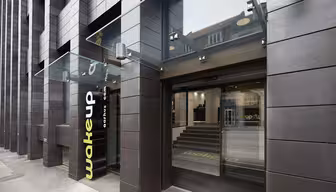 Modern hotellentré med mörk tegelfasad och stor glastak. Gul "wakeup"-logotyp nära de öppna skjutdörrarna i glas. Steg inuti leder till receptionen. Urban miljö synlig genom glasreflektion.