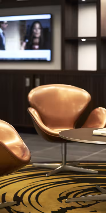 En modern loungedel med orange, svängda stolar och runda bord på en gul och svart mönstrad matta. En väggmonterad TV finns i bakgrunden, placerad i mörka trähyllor.