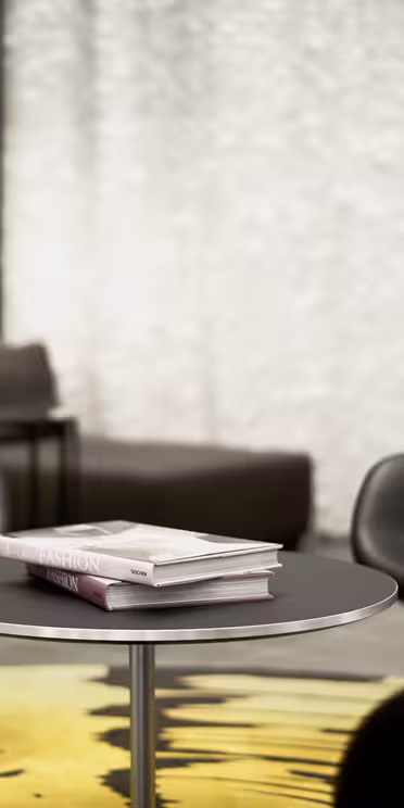 En modern loungedel med tre svarta, svängda stolar runt ett runt bord. Två böcker ligger staplade på bordet. Bakgrunden har strukturerade vita väggar, en mörk soffa och en väggmonterad skärm. En gul och svart matta är delvis synlig.