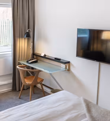 Ett modernt hotellrum med en platt-TV monterad på en vit vägg. Det finns ett skrivbord med en glasskiva, en telefon, en svart lampa och en trästol. Rummet har en säng med vitt täcke och ett fönster med stängda gardiner. En blå stol är delvis synlig.