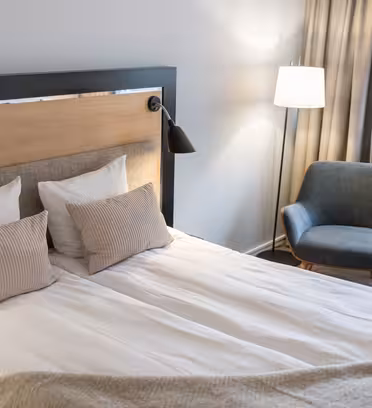 Ett snyggt arrangerat sovrum med en stor säng med beige och vita kuddar och en grå filt. En modern läslampa är fäst på sänggaveln. I närheten står en blå stoppad stol vid en golvlampa och ett fönster med gardiner.
