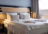 En snyggt bäddad hotellsäng med fräscha vita sängkläder och mjuka beiga kuddar. Två kläder läggs på sängen. Svarta vägglampor lyser upp sänggaveln i trä. Ett stort fönster med beiga gardiner finns till höger.
