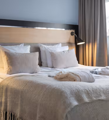 En snyggt bäddad hotellsäng med fräscha vita sängkläder och mjuka beiga kuddar. Två kläder läggs på sängen. Svarta vägglampor lyser upp sänggaveln i trä. Ett stort fönster med beiga gardiner finns till höger.