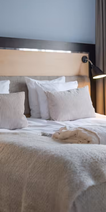 En snyggt bäddad hotellsäng med fräscha vita sängkläder och mjuka beiga kuddar. Två kläder läggs på sängen. Svarta vägglampor lyser upp sänggaveln i trä. Ett stort fönster med beiga gardiner finns till höger.