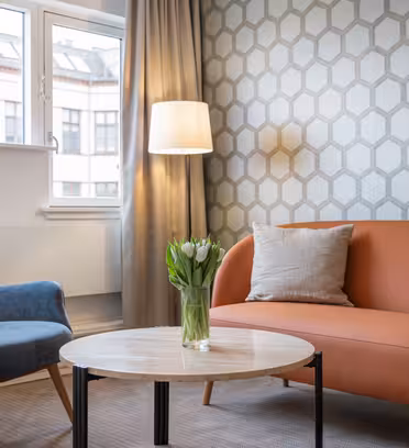 Ett mysigt vardagsrum med en blå fåtölj och en orange soffa som omger ett runt soffbord i trä. En vas med vita tulpaner står på bordet. Väggarna har ett geometriskt mönster och ett fönster med gardiner och en golvlampa ger ljus.