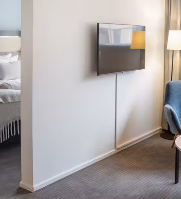 Ett modernt hotellrum har en mysig sittgrupp med en blå fåtölj och ett runt bord med en vas med tulpaner. En platt-TV är monterad på väggen. Sovdelen, delvis synlig, har en prydligt bäddad säng med kuddar och en beige säng.