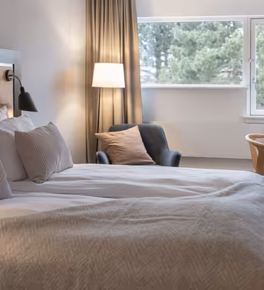 Ett mysigt hotellrum med en stor säng klädd i vita sängkläder och beiga kuddar. En fåtölj, lampa och ett litet skrivbord med en stol finns nära fönstret. Neutrala toner skapar en lugn atmosfär och en skogsutsikt syns genom fönstret.