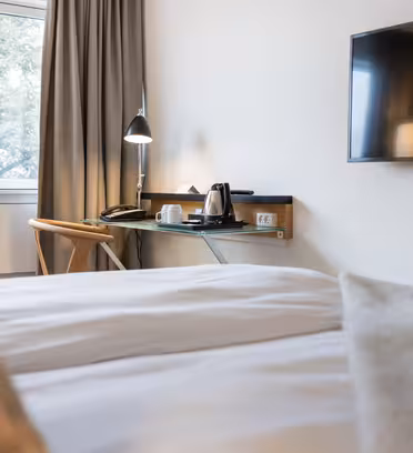 Ett mysigt hotellrum med en snyggt bäddad säng i förgrunden. Ett skrivbord med en lampa, en stol och en bricka med en vattenkokare och koppar står vid fönstret. En TV är monterad på intilliggande vägg. Naturligt ljus filtrerar genom fönstret och lyser upp utrymmet.