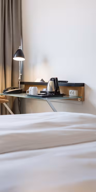 Ett mysigt hotellrum med en snyggt bäddad säng i förgrunden. Ett skrivbord med en lampa, en stol och en bricka med en vattenkokare och koppar står vid fönstret. En TV är monterad på intilliggande vägg. Naturligt ljus filtrerar genom fönstret och lyser upp utrymmet.