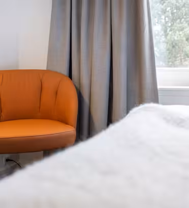 Myshörna med en orange dämpad stol framför grå gardiner och ett fönster. Vitt överkast i förgrunden tipsar om en bekväm, minimalistisk sovrumsmiljö.
