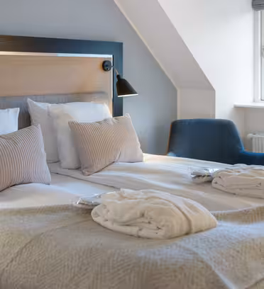 Ett mysigt hotellrum med en snyggt bäddad dubbelsäng med två vita badrockar och kuddar. En blå fåtölj är nära ett fönster, vilket låter naturligt ljus lysa upp rummet, som har modern, minimalistisk inredning.