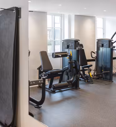 Ett modernt gym med träningsutrustning, inklusive elliptiska maskiner, en benpress och andra träningsmaskiner. Rummet har ljusa väggar, stora fönster och en spegelvägg som reflekterar utrymmet. Golvet är ett mörkt, strukturerat material.