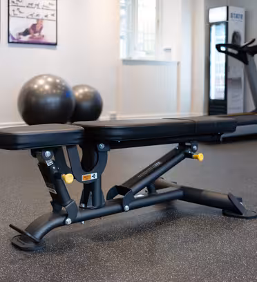 En gymmiljö visar upp en svart justerbar bänk i förgrunden. Bakom den finns träningsbollar, ett löpband och instruktionsaffischer på väggen. Gymgolvet är täckt med en spräcklig gummimatta.