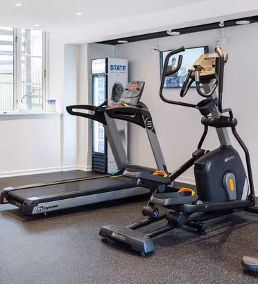 Ett litet gym med löpband, elliptisk maskin, stationär cykel och träningsboll på ett mörkt gummigolv. Det finns ett fönster som släpper in naturligt ljus och en vätskestation med en TV-skärm monterad på väggen.