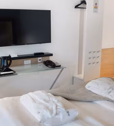 Hotellrum med en väggmonterad TV, skrivbord med kaffebryggare och koppar, lampa, telefon och snacks. En säng står i förgrunden med vikta kläder. En klädhängare finns på väggen och en liten trähylla nära dörren. Vita väggar och belysning förstärker utrymmet.