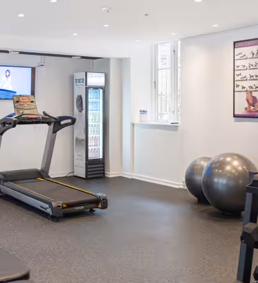 Ett modernt gym med träningsutrustning inklusive ett löpband, stationär cykel, hantlar, träningsbollar och en bänk. En TV-skärm visar ett program och träningsaffischer finns på väggen. Ett fönster släpper in naturligt ljus.
