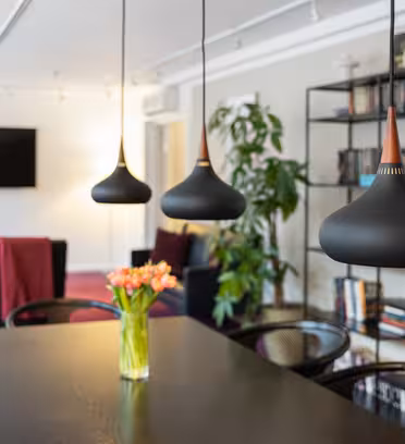Ett modernt vardagsrum med tre svarta taklampor hängande ovanför ett matbord. En vas med orange tulpaner står på bordet. I bakgrunden finns en bokhylla, en platt-TV och röddämpade stolar.