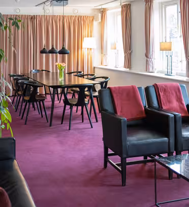 Ett modernt rum med en magentafärgad matta har ett långt bord omgivet av svarta stolar. Svarta lädersoffor med röda kuddar vetter mot ett soffbord i marmor. Fönster från golv till tak har mjukt rosa gardiner, och det finns krukväxter och hyllor i bakgrunden.