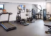 Ett modernt gym med olika träningsredskap, inklusive ett löpband, elliptisk, stationär cykel och viktmaskiner. Rummet har en platt-TV på väggen och stark belysning. Golvet är täckt med gymgolv i gummi.