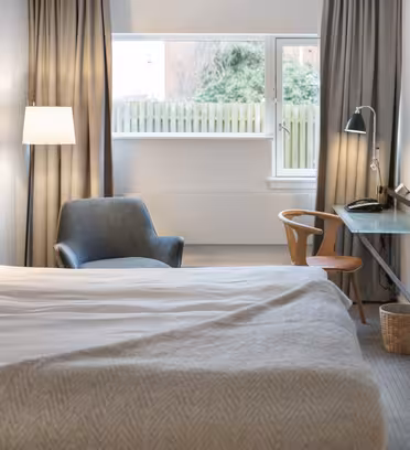 Ett modernt hotellrum med en snyggt bäddad säng, grå filt och vit kudde. Det finns en grå fåtölj, en väggmonterad TV, ett litet skrivbord med en stol, en lampa och ett stort fönster med gardiner som släpper in naturligt ljus.