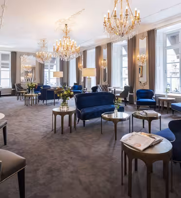 Elegant lounge med blå sammetsstolar, marmorbord och utsmyckade ljuskronor. Stora fönster med grå gardiner släpper in naturligt ljus. Dekorativa speglar och blomsterarrangemang förstärker den lyxiga atmosfären. Tidningar ligger på ett bord.