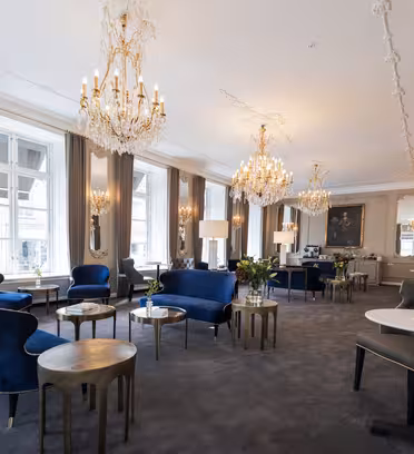 Lyxigt rum med utsmyckade ljuskronor, sittplatser i blå sammet och bord med guldaccent. Stora fönster släpper in naturligt ljus och eleganta speglar förstärker rymligheten. Inredningen kombinerar klassiska och moderna element i en sofistikerad miljö.