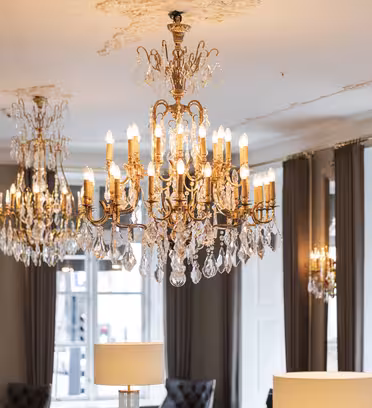 Elegant rum med utsmyckade kristallkronor som hänger i taket och reflekterar ljus. Stora fönster med gardiner syns. Ljuskronorna tillför en lyxig atmosfär till utrymmet.