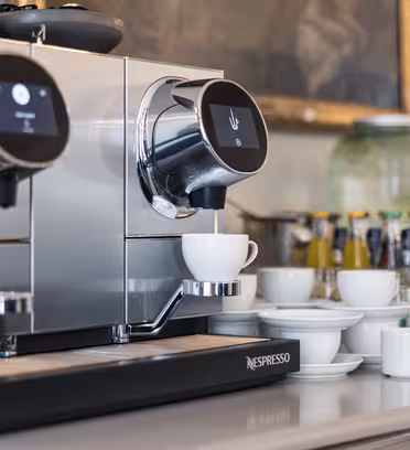 En elegant silver kaffemaskin med två dispensrar, en fyller en vit kopp placerad under. I närheten syns prydligt staplade vita koppar och fat, olika dryckesflaskor och två stora vattenautomater i bakgrunden.