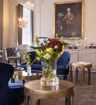 Elegant lounge med blå sammetsstolar, gyllene bord och ett blommigt mittpunkt i en vas. Ett stort porträtt hänger på väggen och ljuset strömmar in genom höga fönster, lysande ljuskronor och dekorativa speglar.