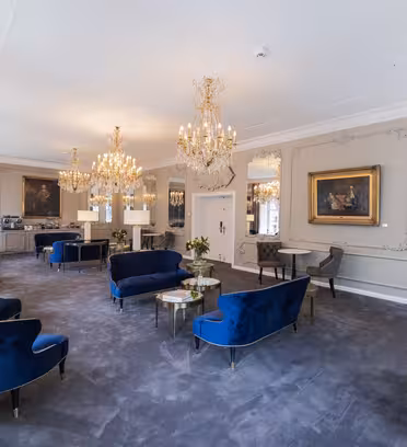 Elegant lounge med plyschblå stolar, utsmyckade ljuskronor och klassiska vägglister. Stora speglar och inramade konstverk pryder de ljusgrå väggarna. Bord med små blomsterarrangemang ger en touch av färg till den överdådiga miljön.
