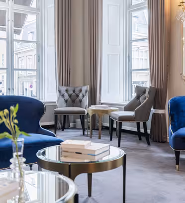 Elegant lounge med blå sammetssoffor, grå stoppade stolar och runda soffbord i glas. Stora fönster med ljusridåer släpper in naturligt ljus som reflekteras på en ljuskrona och spegel. En hög med böcker och en grön växt pryder borden.
