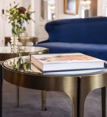 Ett sofistikerat vardagsrum har en mörkblå sammetssoffa och eleganta runda soffbord i metall med glasskivor. En dekorativ bok vilar på det ena bordet, och en vas med blommor pryder det andra. Rummet har en plyschgrå matta.