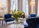 En lyxig lounge med blå sammetsstolar och runda träbord. En glasvas med diverse blommor sitter på ett bord i förgrunden. Eleganta ljuskronor och stora fönster med grå gardiner pryder det ljusa utrymmet.