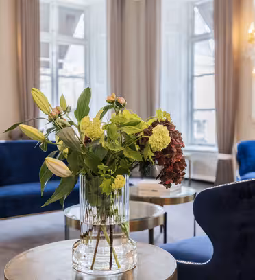 En lyxig lounge med blå sammetsstolar och runda träbord. En glasvas med diverse blommor sitter på ett bord i förgrunden. Eleganta ljuskronor och stora fönster med grå gardiner pryder det ljusa utrymmet.