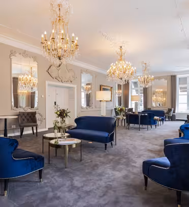 Elegant rum med plyschblå sammetssoffor och fåtöljer. Stora utsmyckade speglar och ljuskronor hänger på väggar och tak. Mjuk belysning och dekorativa accenter skapar en lyxig atmosfär, med små runda bord placerade mellan sittplatserna.