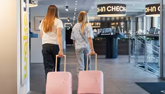 Två personer med långt hår som drar i rosa resväskor går mot ett incheckningsområde på hotellet. Interiören är modern, med stark belysning och tydlig skyltning. Personerna bär vardagskläder, inklusive jeans och t-shirts.