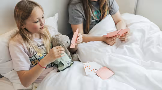 Två unga flickor sitter på en säng med vita lakan och spelar kort. En flicka håller i en fylld leksak och snackspåse, medan den andra tittar på sina kort. Solljuset strömmar in från fönstret bakom dem.