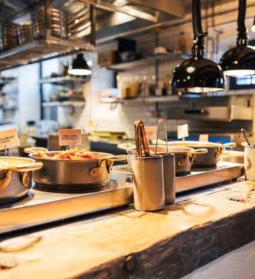 En rustik restaurangköksbänk med krukor med mat på en träyta. Varje kruka har en liten skylt som anger dess innehåll. Taklampor lyser upp området och bakgrunden visar ett hektiskt kök med hyllor och redskap.