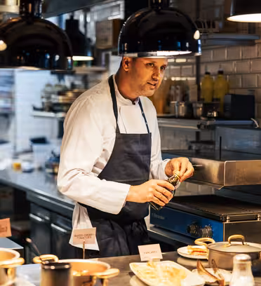 En kock i vit jacka och blått förkläde smaksätter mat med kryddor i ett storkök. Bänkskivan är fylld med krukor och mat, och hyllor med kryddor och ingredienser syns i bakgrunden. Varm belysning lyser upp scenen.