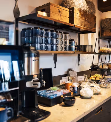 En mysig cafédisk med kaffebryggare, muggar staplade på hyllor, en mängd olika snacks och trälådor. Disken har korgar med bakverk, glasburkar och färsk frukt, vilket skapar en varm och välkomnande atmosfär.