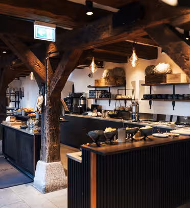 Rustik restauranginteriör med träbjälkar och modern belysning. Borden är dukade med tallrikar och redskap, och hyllor rymmer keramik och dekor. Atmosfären är mysig och välkomnande, med en blandning av naturliga trä- och stenstrukturer.